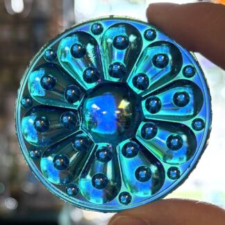 Close-up of an iridescent blue-green round disk with petal-like segments and small raised dots, held between two fingers.