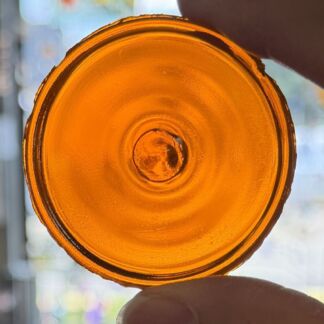 Close-up of an amber glass bottle cap held by fingers, showing the circular rim and inner core against a bright window light.